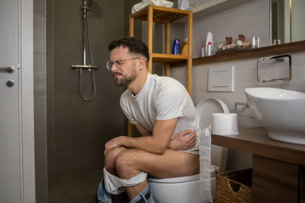 A man with glasses sitting on a toilet in a modern bathroom, holding his stomach with a pained facial expression, indicating digestive distress or constipation.