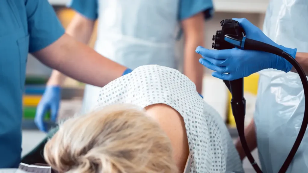 A medical team in blue scrubs and gloves assists a patient wearing a hospital gown during a procedure with an colonoscope in a clinical setting.